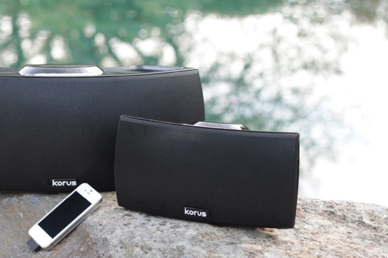 Two Korus speakers sitting on a rock next to a body of water.