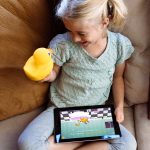 A girl sitting on a couch with Edwin The Duck and a tablet.