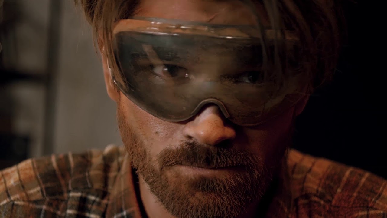 A man wearing goggles and a beard carefully straps in before jumping onto a gravel track.