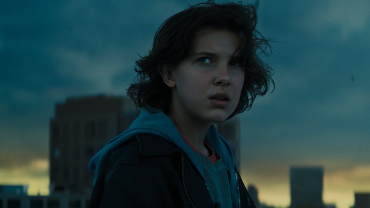 A young girl is standing in front of a city with a cloudy sky in the Official Trailer for "Godzilla, King of Monsters.