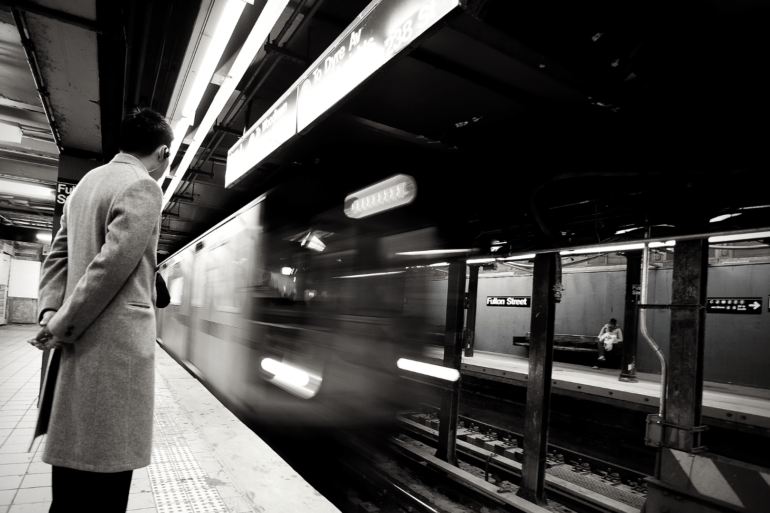 A black and white photo of a man waiting for a train at an MTA station.