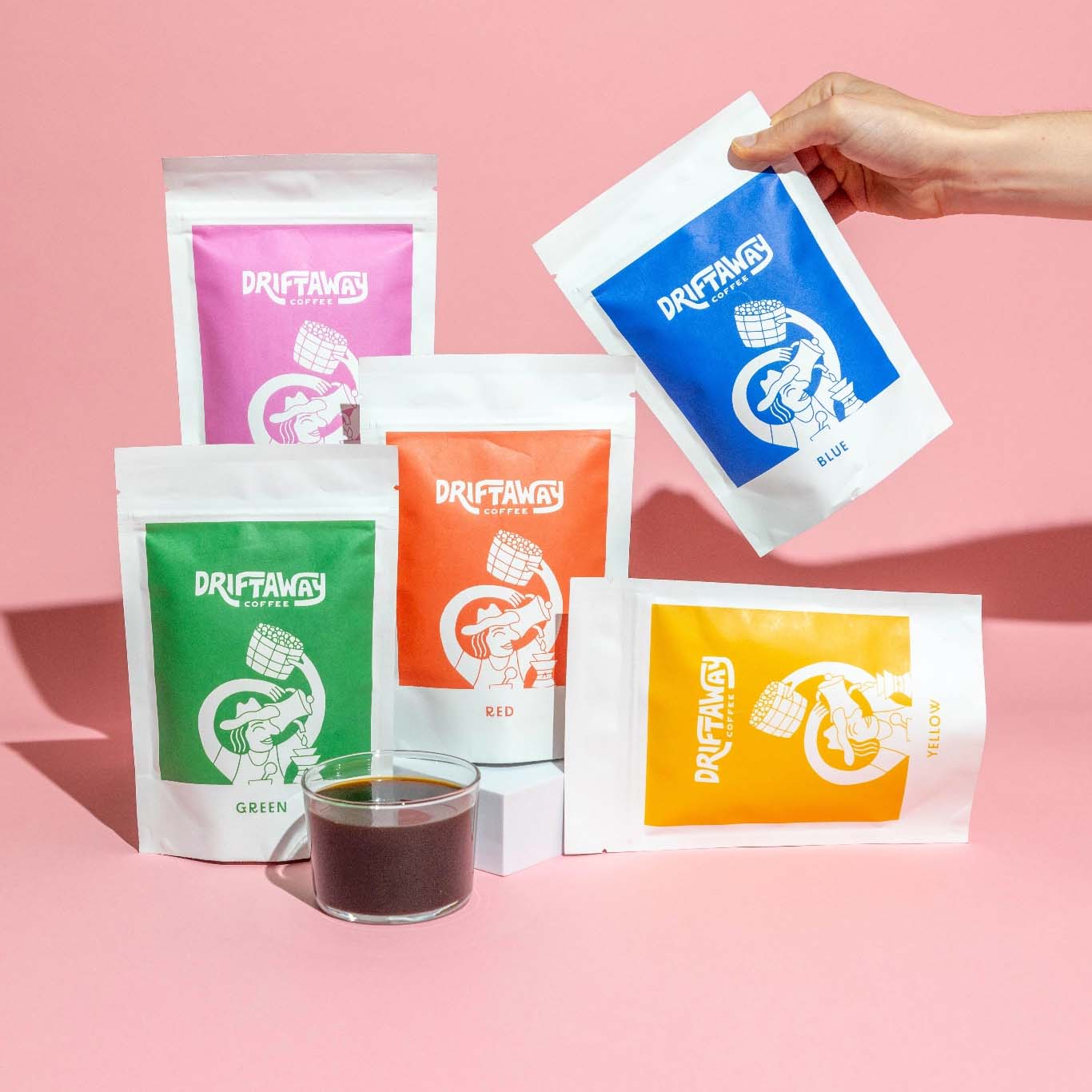 A person holding a bag of coffee and a cup of coffee in front of a pink background.