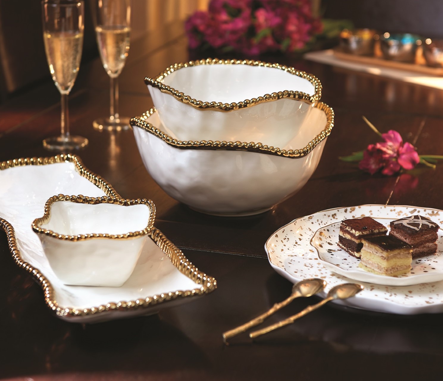 An elegant table setting featuring gold-rimmed ceramic bowls, a rectangle platter, and a dessert plate with layered cake accompanies two champagne flutes on a wooden table—perfect for inspiration in the RAGE Works Holiday Gift Guide.