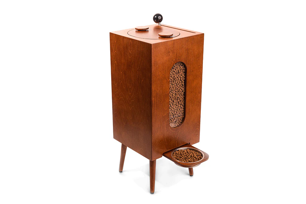 This wooden pet food dispenser, a standout from the Holiday Tech Gift Guide, features a vertical oval window revealing kibble, a lid on top, and a bowl at the bottom on angled legs. It's perfect for adding style to your pet’s dining routine.