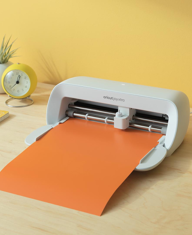 A Cricut Joy Xtra machine on a wooden table holds an orange sheet, with a small clock and a plant in the background against a yellow wall. Perfect for any craft enthusiast, it’s an ideal pick from the Tech Gift Guide curated by RAGE Works.